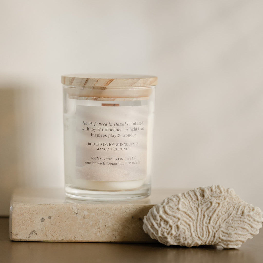 Keiki Candle Mango Sticky Rice in a glass jar with wooden lid alongside decorative coral piece on a natural stone surface.